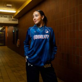 Woman in the blue EQUALITY jersey standing in a dimly lit corridor 